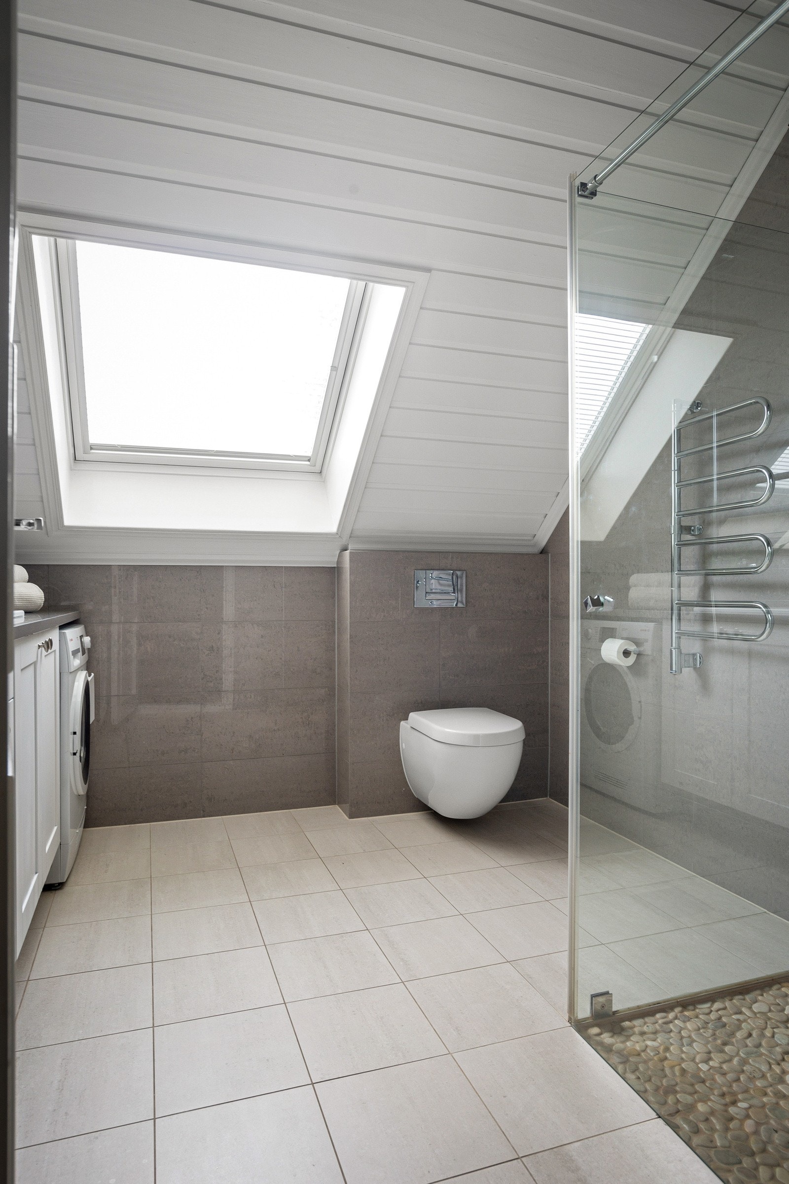 Videre er badet utstyrt med vegghengt toalett og et tidsriktig dusjhjørne med glassvegger. Et stort takvindu slipper inn rikelig med naturlig lys. Galleribilde