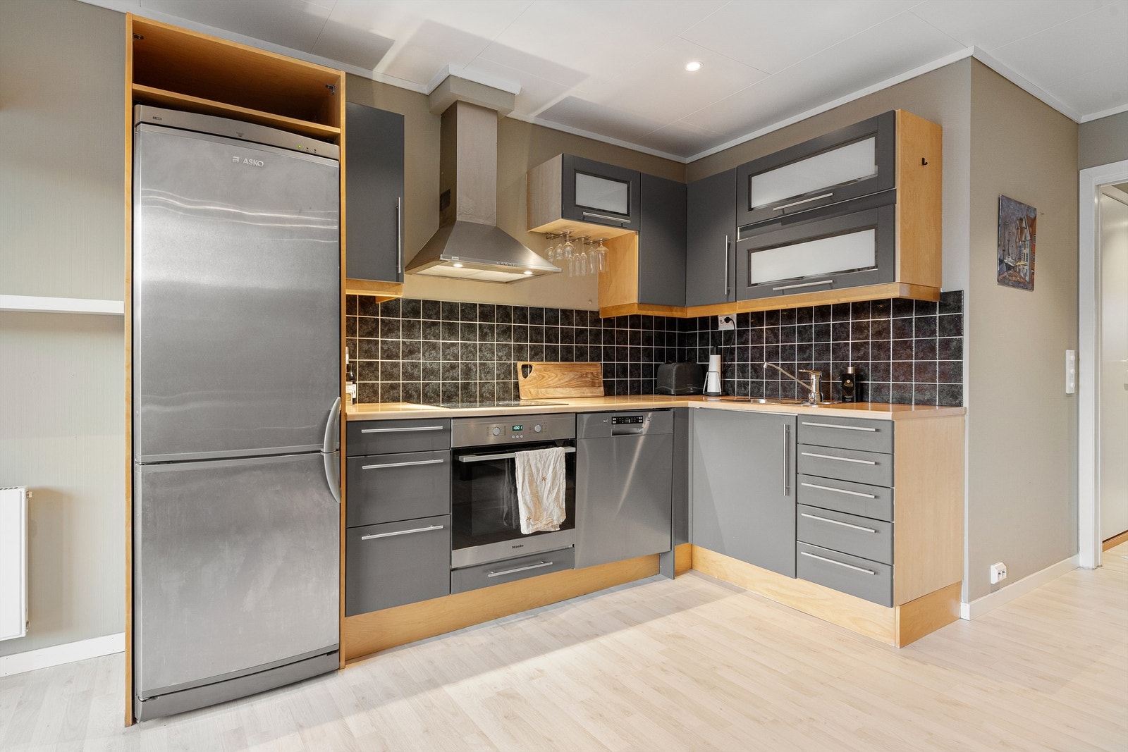 Kjøkkeninnredning med glatte fronter og benkeplate av laminat. Integrert stekovn og platetopp. Frittstående oppvaskmaskin og kjøleskap med fryser Galleribilde