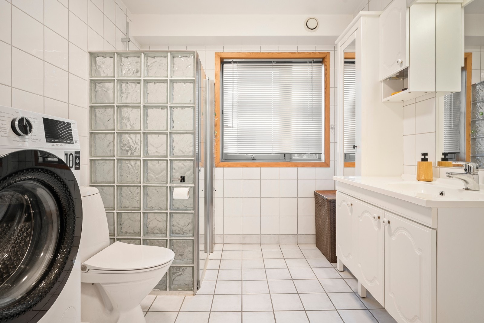 Bad med flislagte overflater på gulv og vegger. Baderomsinnredning med profilerte lyse fronter og overhengende speil med belysning. Dusjen er plassert i hjørnet med glassbyggerstein, glassdør og dusjarmatur med dusjsett. Galleribilde