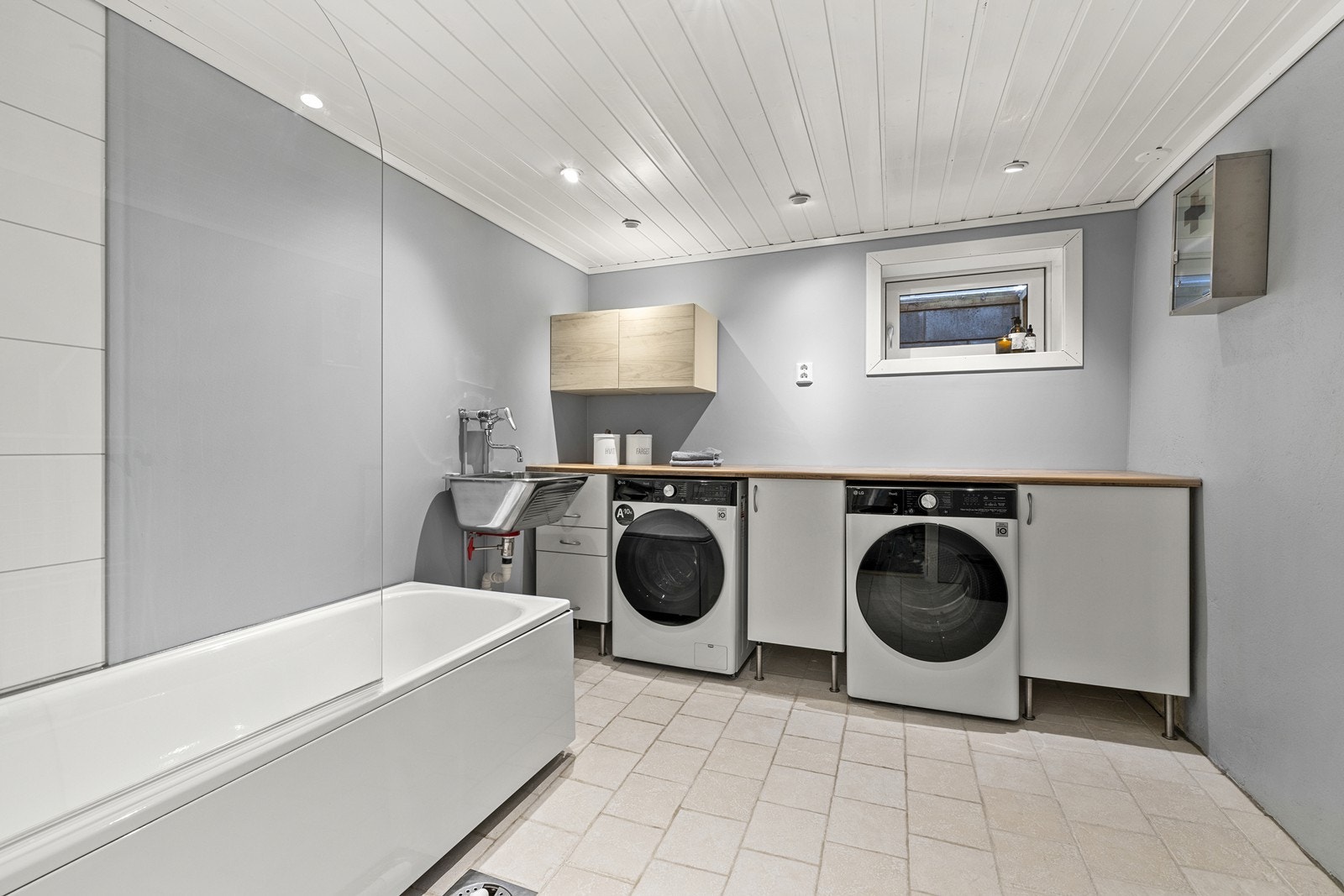 Et stort og praktisk vaskerom med flislagt gulv. Vaskerommet er utstyrt med badekar med dusjløsning, benkeplate med opplegg for vaskemaskin og tørketrommel i underkant, samt utslagsvask. Varmekabler i gulv og downlights i tak. Galleribilde