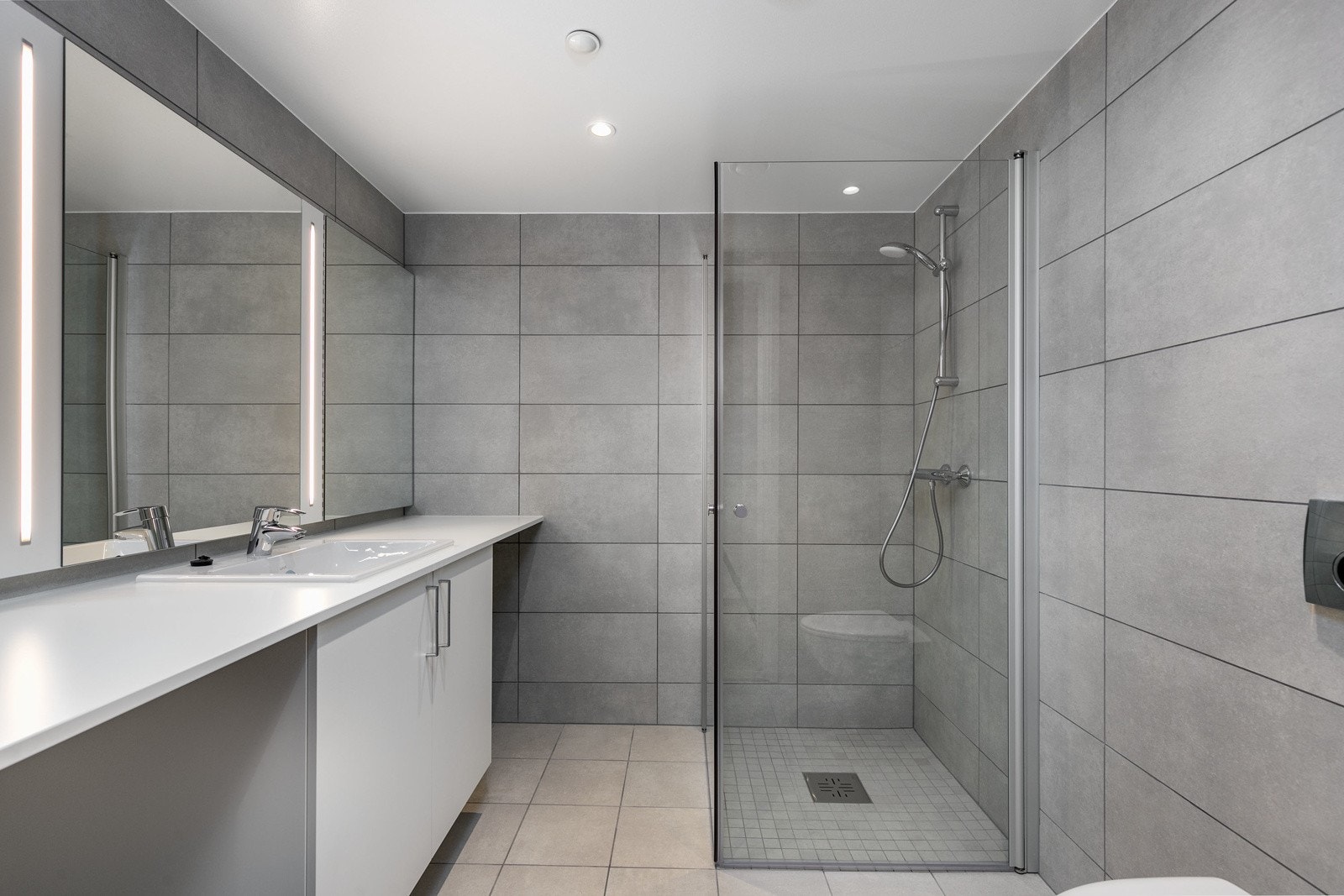 Delikat flislagt bad med vegghengt toalett, dusjsone med innfellbare dører samt servant med skapinnredning. Galleribilde