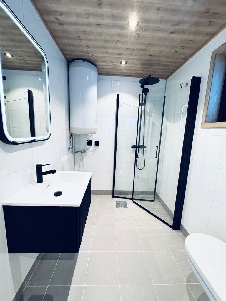 Bad leveres med flis på gulv med varme og stilige baderomsplater på vegg. Opplegg for vaskemaskin, innfellbart dusjhjørne og baderomsinnredning med speil Galleribilde