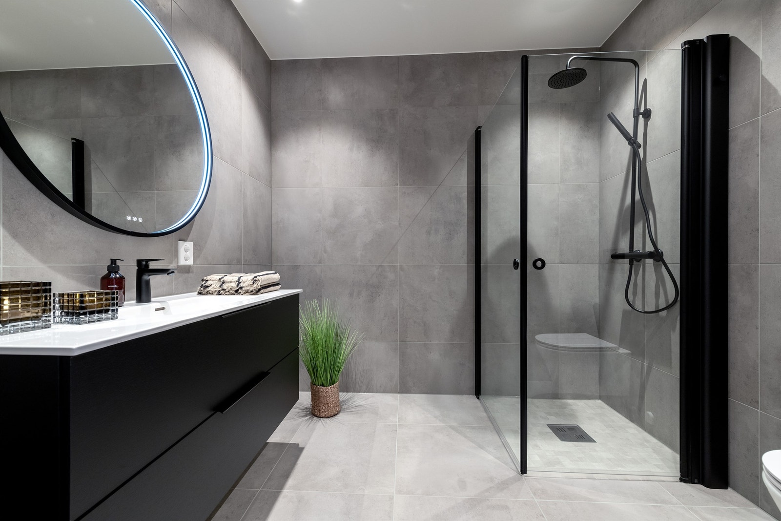 Lekkert flislagt bad med servantinnredning, speil med lys, dusjhjørne med glassvegger og vegghengt dusjarmatur Galleribilde