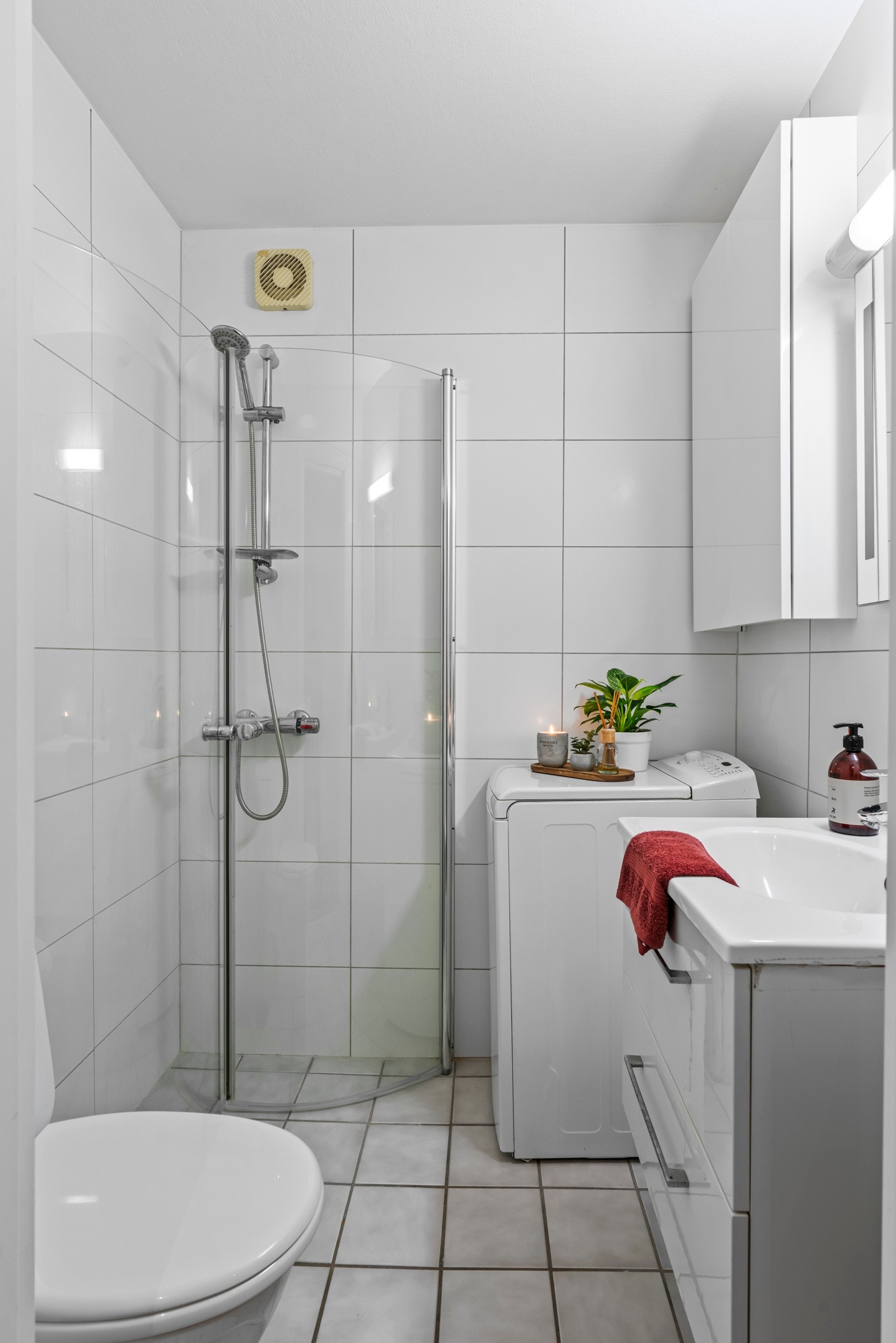 Badet i leiligheten er tidsløst med lyse fliser og varmekabler med termostat i badegulvet. Her er det opplegg for vaskemaskin, gulvmontert toalett og dusjhjørne med buet front. Galleribilde