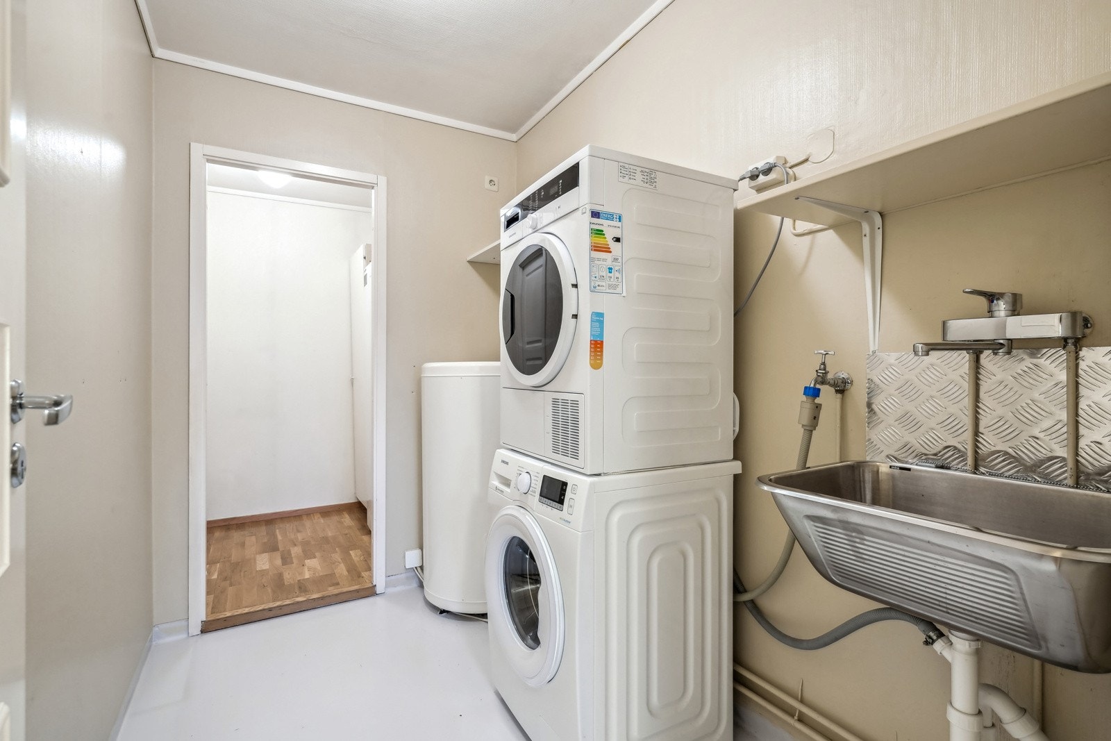 Praktisk vaskerom med opplegg for vaskemaskin og tørketrommel. Rommet har en stor utslagsvask med hylle over. Galleribilde