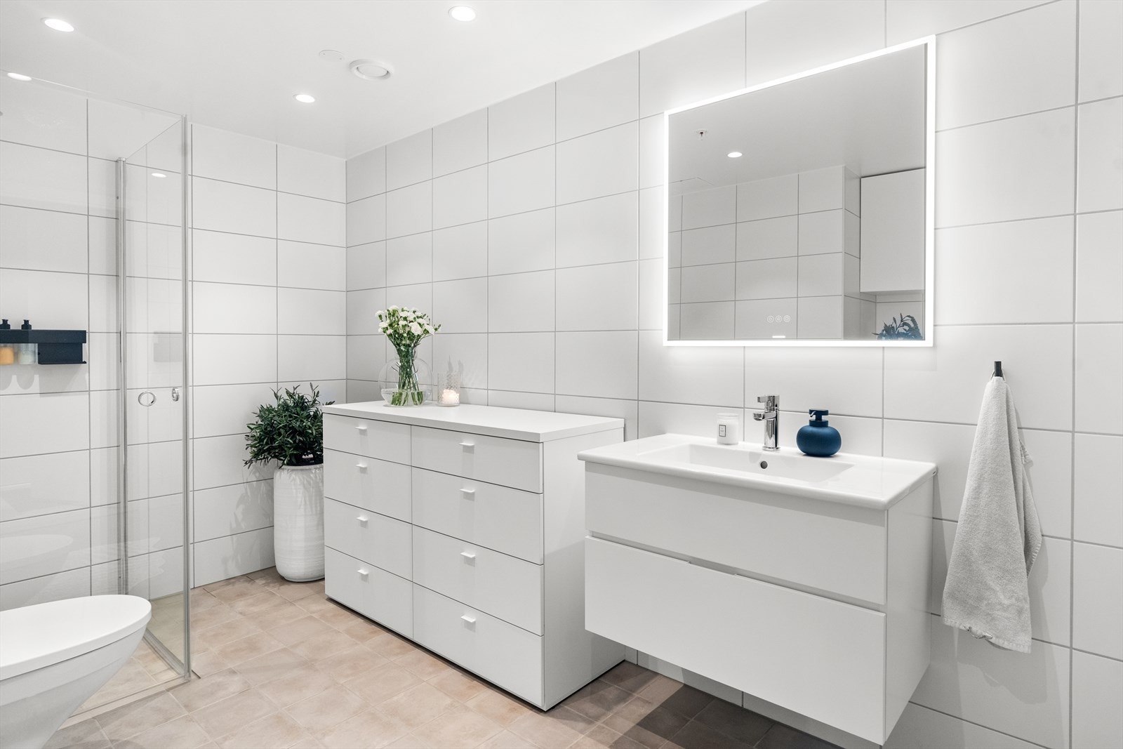 Videre er badet utstyrt med innredning bestående av skap med glatte fronter, servant og speil med lys. Det er også satt inn dusj med glassdører, veggfestet klosett og opplegg for vaskemaskin. Galleribilde