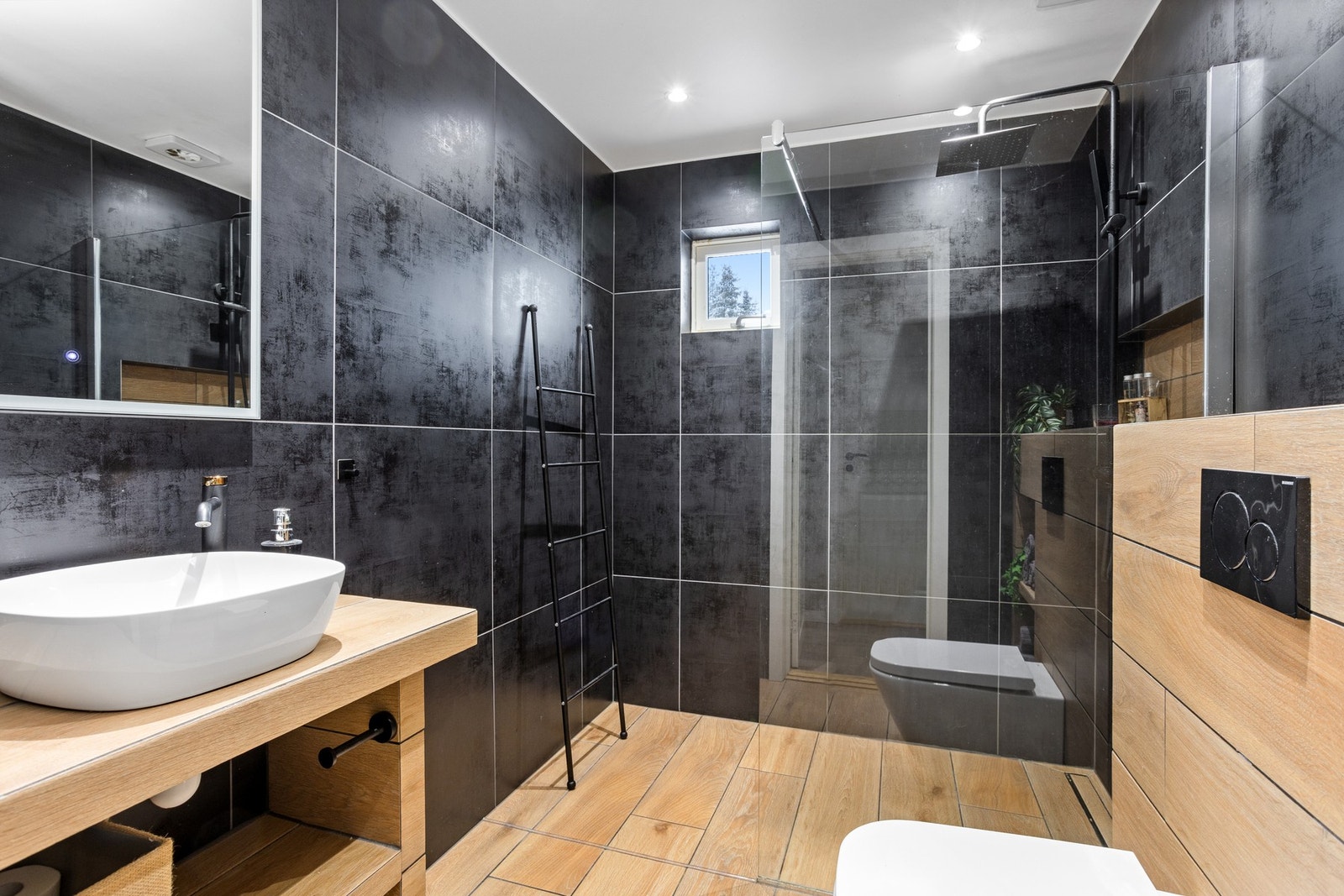 Delikat baderom med fliser på vegger og gulv, spesiallaget servantbenk, vegghengt toalett og dusjhjørne med regnfallsarmatur. Galleribilde