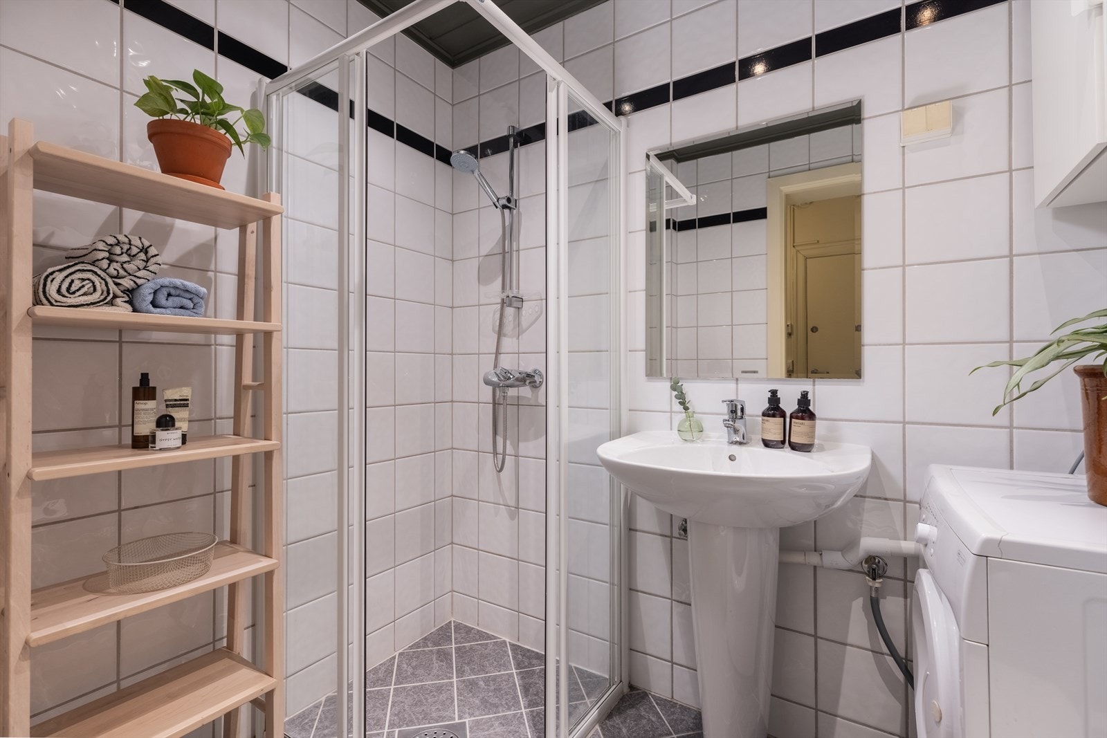 Lyst flislagt baderom med en innredning bestående av gulvmontert toalett, servant, speil, veggskap, dusjnisje med skyvedører. Opplegg for vaskemaskin. Galleribilde