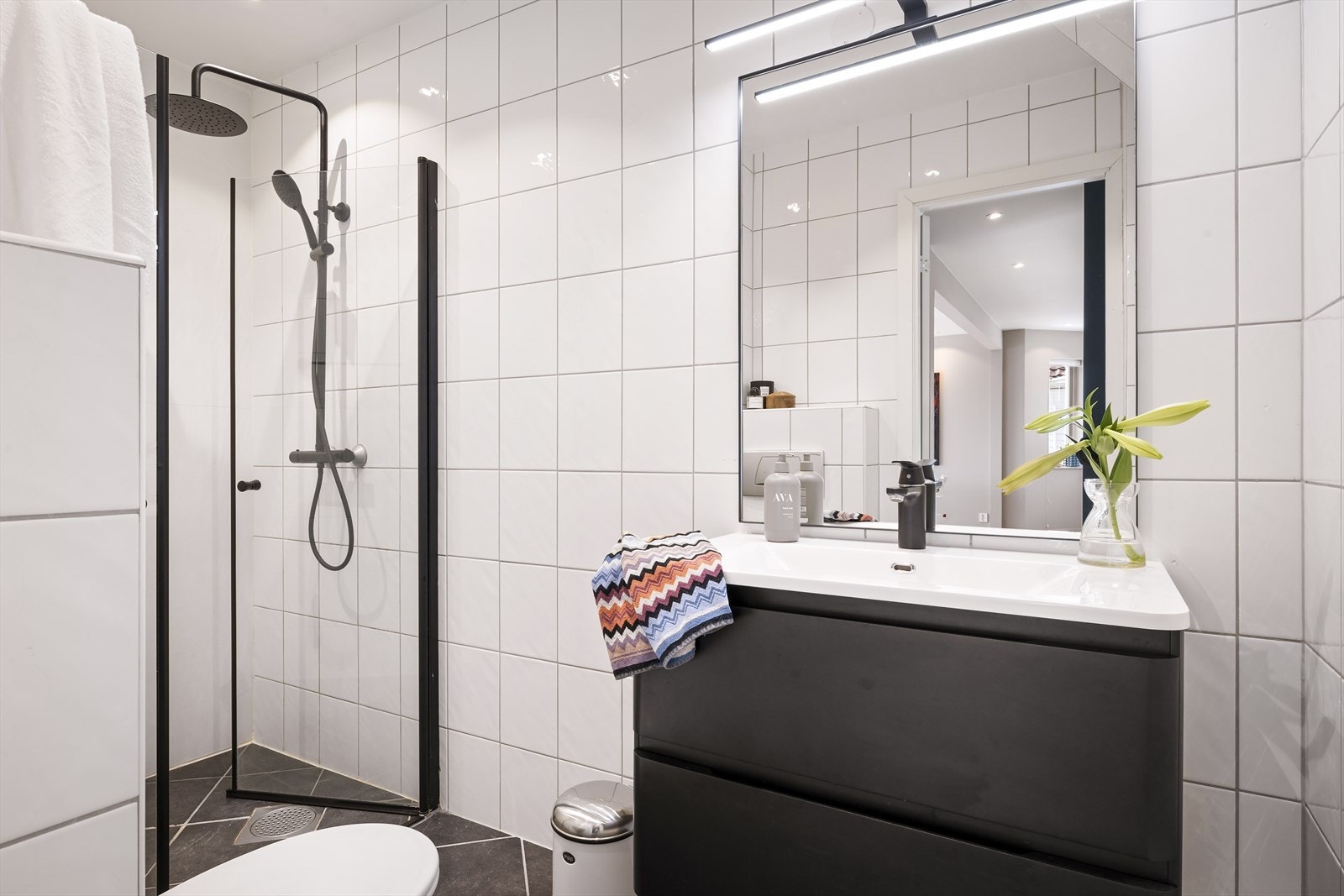 Badet er pent helfliset i en klassisk sort-hvit stil og er innredet med servant i møblement, vegghengt toalett, speil og belysning, dusjhjørne med dører i glass. Galleribilde