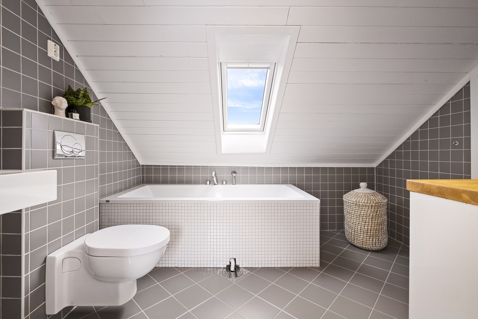 Badet er helfliset i en delikat gråfarge og utstyrt med badekar, servant, vegghengt toalett og vegghengt skap Galleribilde