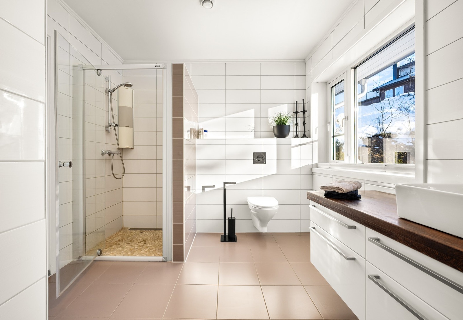 Med tiltalende og tidløse fliser på gulv og vegger, doble servanter i innredning med massiv heltre benkeplate, veggmontert toalett og dusjhjørne Galleribilde