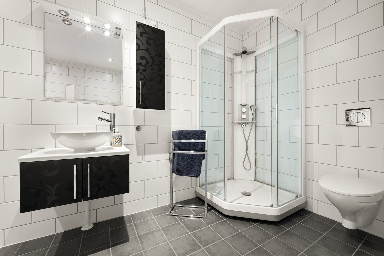 Bad/WC/vaskerom med flislagte gulv og vegger. Dusjkabinett, veggmontert toalett og servant i innredning. Galleribilde