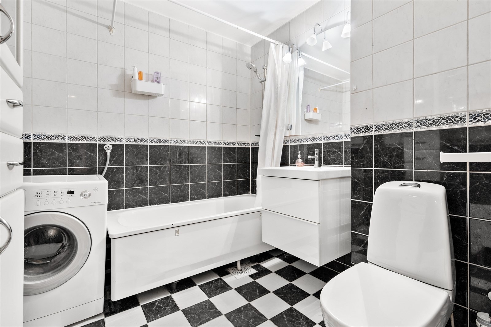 Eldre flislagt bad innredet med frittstående toalett, badekar med dusjarmatur og forheng, servant med underskap og speil med lys over. Galleribilde