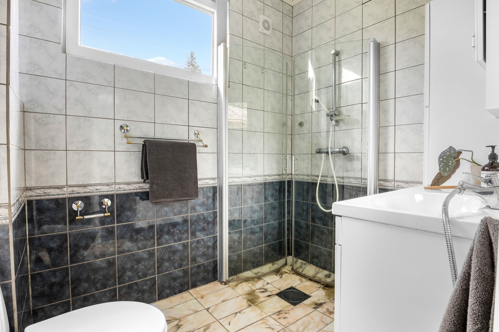 Badet i første etasje har fliser på gulv og vegger, vindu og elektrisk avtrekksvifte. Rommet er utstyrt med dusjhjørne med innsvingbare glassdører, et gulvstående toalett og en hvit baderomsinnredning med servant Galleribilde