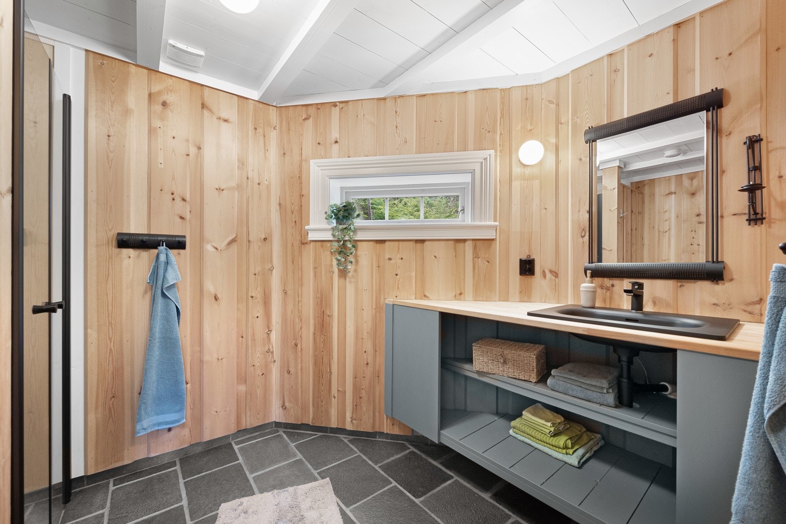 Hyttens baderom har fliser på gulv, panel på vegger og malt tak. Det er en plassbygget baderomsmøbel med slette fronter, med hylle, malt i en flott farge. Galleribilde