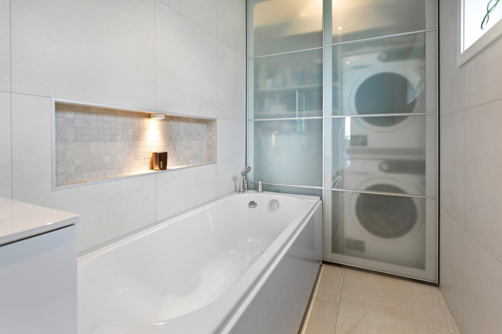 Bad/vaskerom med fliser og varmekabler på gulv, flis på vegger. Badekar, veggmontert toalett, servant med underskap, vv-bereder og ventilasjon i himling. Det er sluk under badekar. Vv-bereder og opplegg for vaskemaskin bak skyvedør Galleribilde