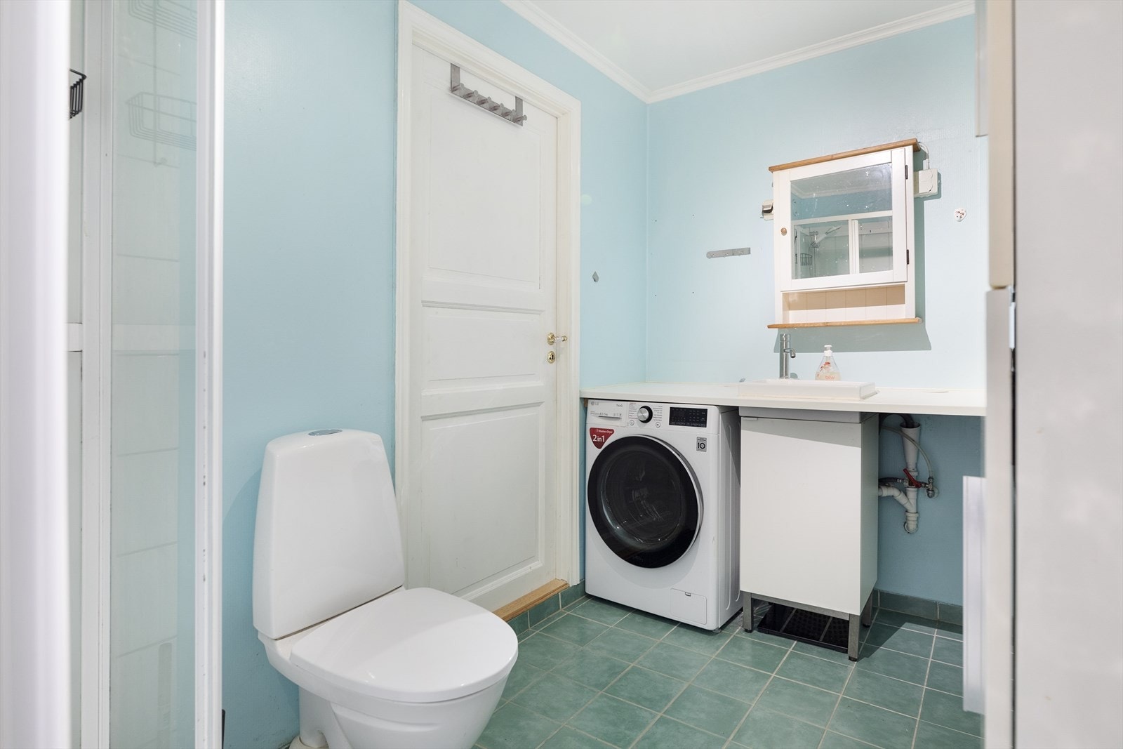 Badet består av laminat benkeplate med nedfelt servant, dusjkabinett, gulvmontert toalett, overskap med speilfront og hylle, og opplegg for vaskemaskin Galleribilde