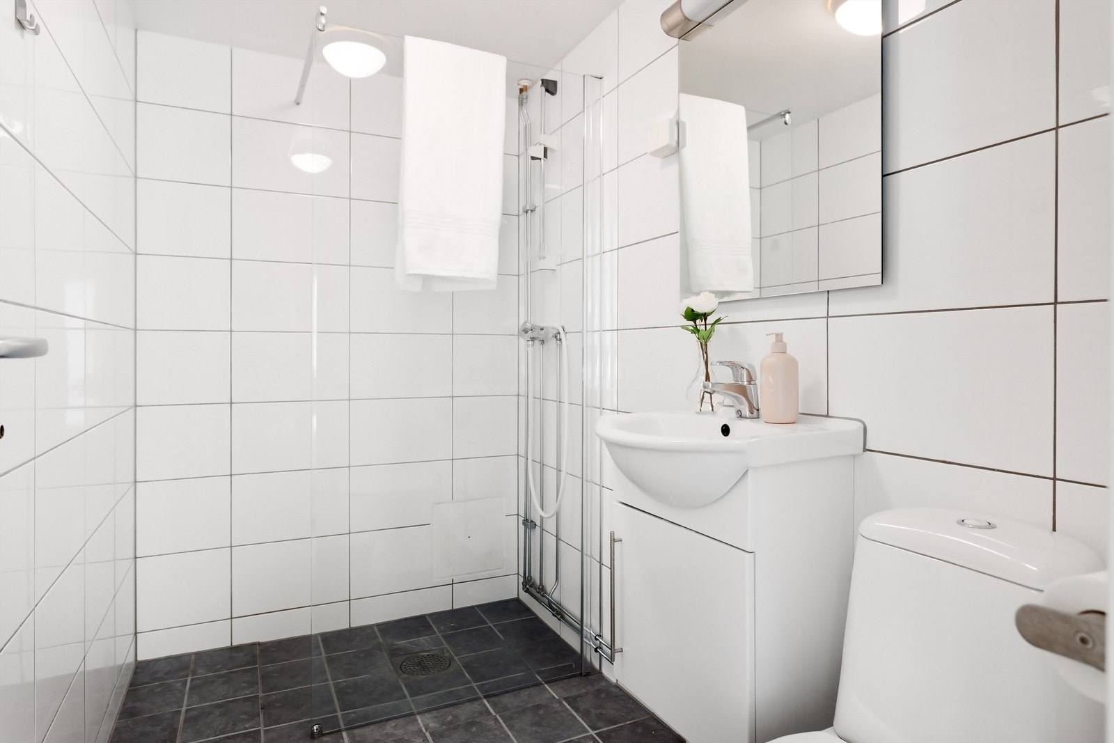 Pent baderom fra 2013 med fliser på gulv med gulvvarme, fliser på vegger og malt himling. Badet har toalett, servant med underskap, dusjnisje med glassvegg og opplegg for vaskemaskin Galleribilde