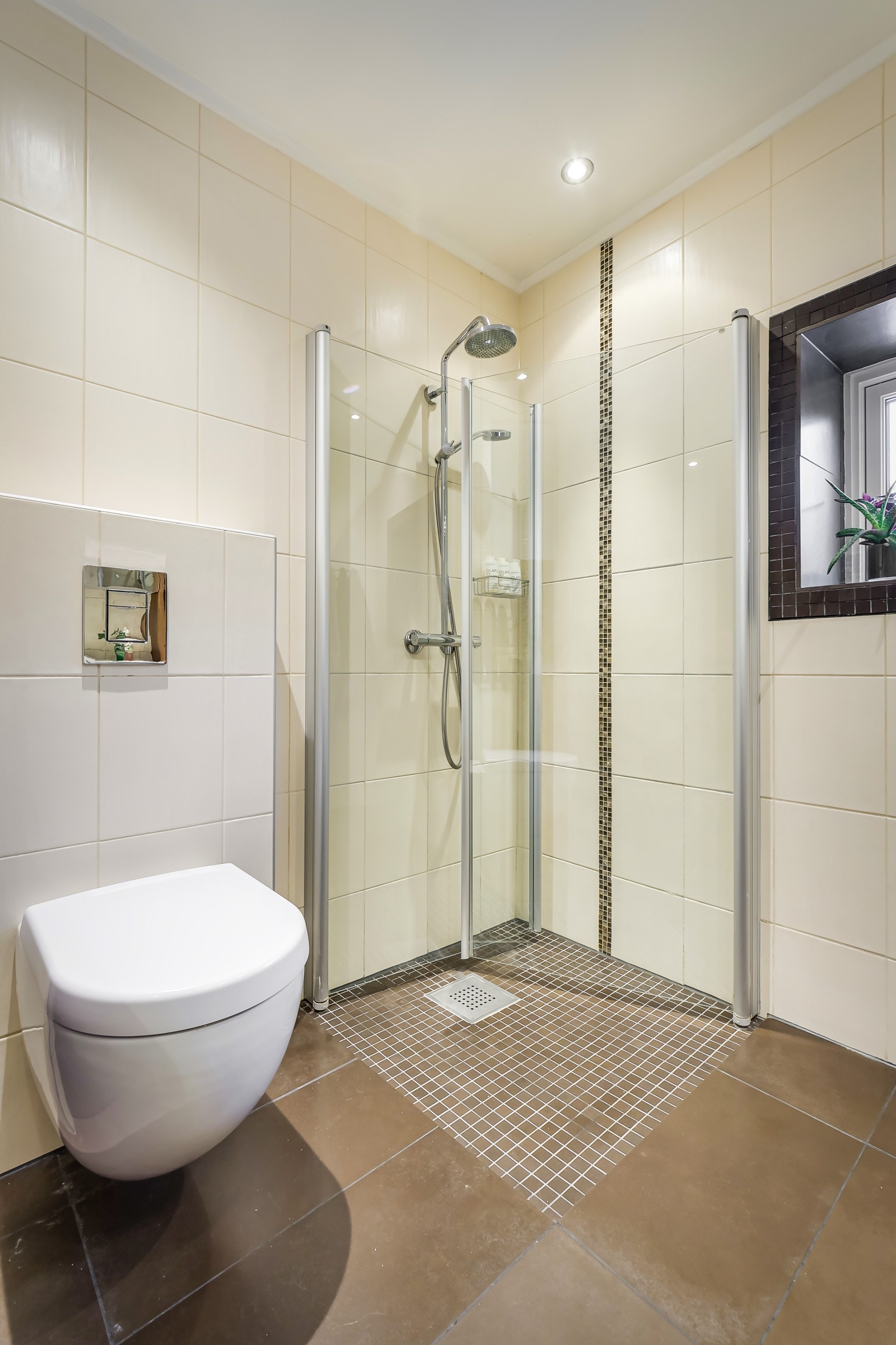 Dusjhjørne med mosaikkfliser på gulv, innfellbare glassdører og både hånd- og regnfallsdusj. Baderommet har også vegghengt toalett. Galleribilde