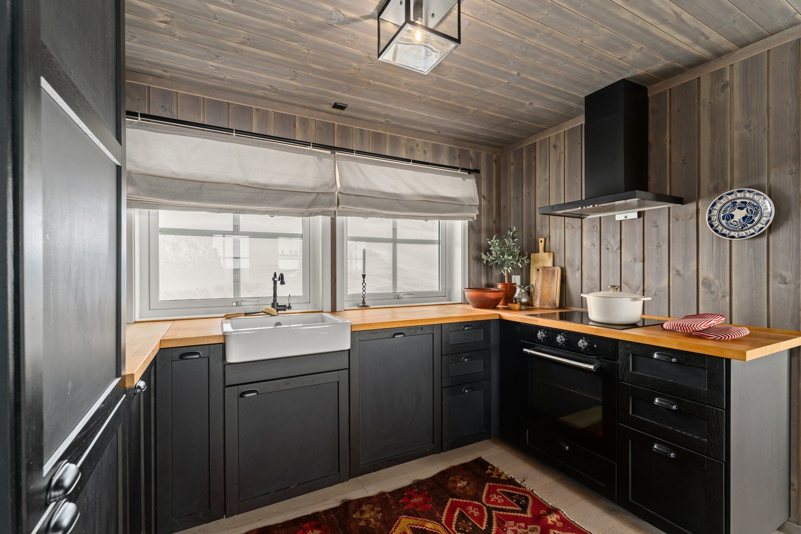 Kjøkkenet har mørke skapfronter, trebenkeplater og en rustikk stil. Store vinduer gir godt lysinnslipp. Veggene og taket er kledd i trepanel. Det er en praktisk vask og en moderne ventilator over koketoppen. Galleribilde