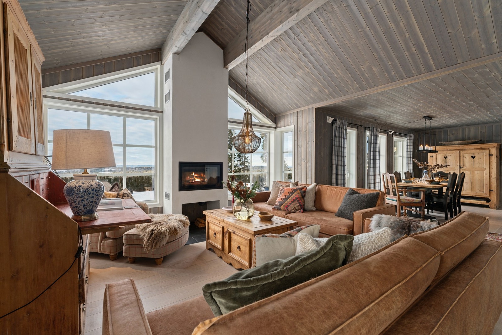 Romslig stue med store vindusflater som gir godt lysinnslipp og utsikt. Peis i midten av rommet gir en lun atmosfære. Åpen løsning mot spiseplass og kjøkken. Tregulv og panelte vegger i naturlige toner. Galleribilde