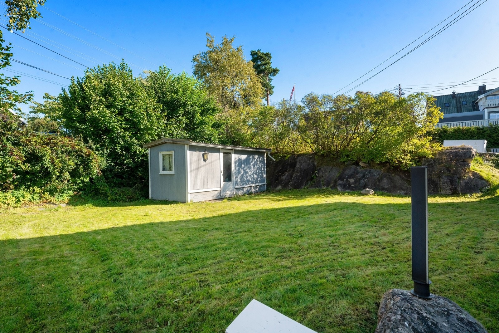 Fra gårdsplassen er det trapp opp til sydvendt plenarealer mellom husveggen og dobbelgarasjen, som kantes av fine prydbusker og rhododendron i grensen mot naboen. Galleribilde