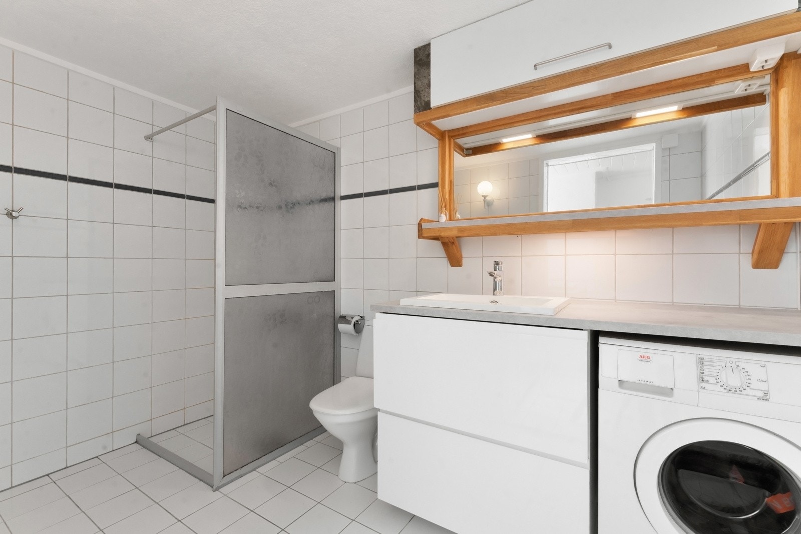 Badet i praktikantdelen har lyse fliser på gulv og vegger, gulvvarme og en dusjnisje med glassvegg. Det er opplegg for vaskemaskin her. Galleribilde