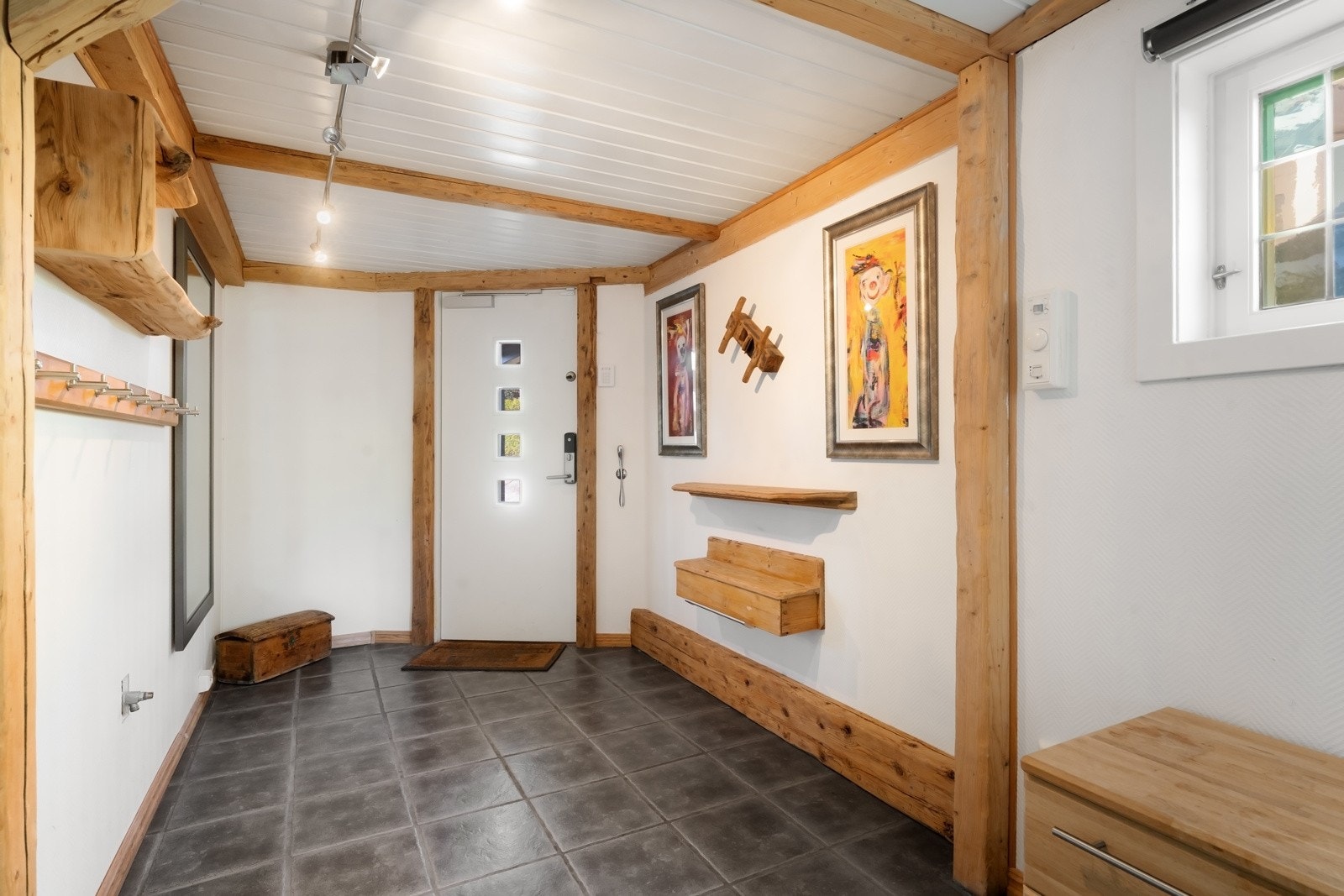 Ytterdøren har praktisk kodelås og fører inn i en romslig hall med mørke gulvfliser, gulvvarme og lysmalte vegg- og takflater, rammet inn av rustikke bjelker. Galleribilde