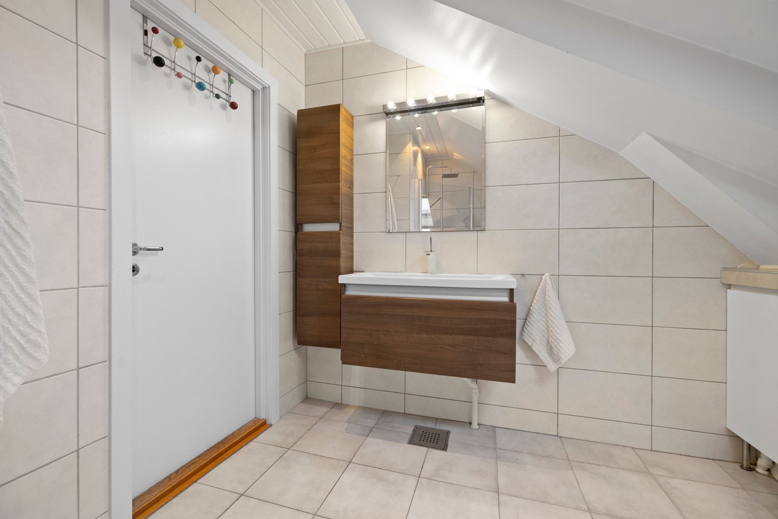 Baderomsinnredningen består av servantskap med glatte, folierte fronter og heldekkende servant. Speil og lysarmatur på vegg over servant. Dusjhjørne med svingdører, sluk i sone, regnsfalldusj og termostatstyrt blandebatteri. Veggmontert toalett. Galleribilde