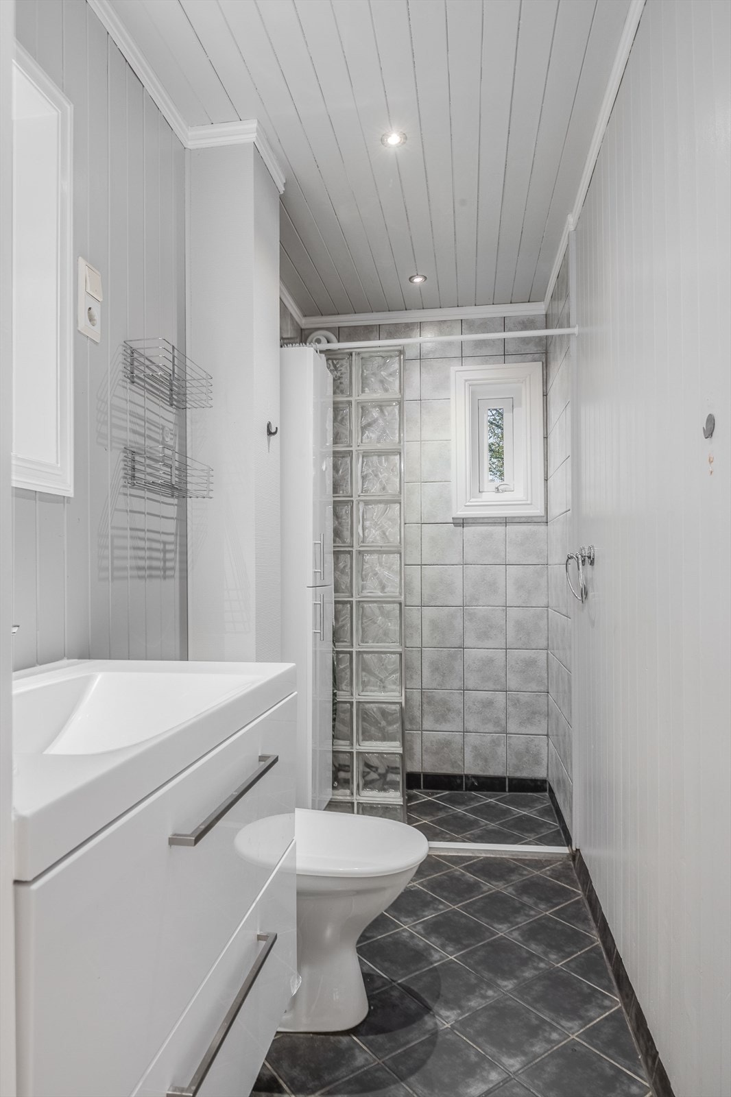 Badet er praktisk plassert i gangen, med flislagt gulv og lysmalt panel. Dusjnisjen har halvvegg av glassbyggerstein og dusjforheng. Galleribilde