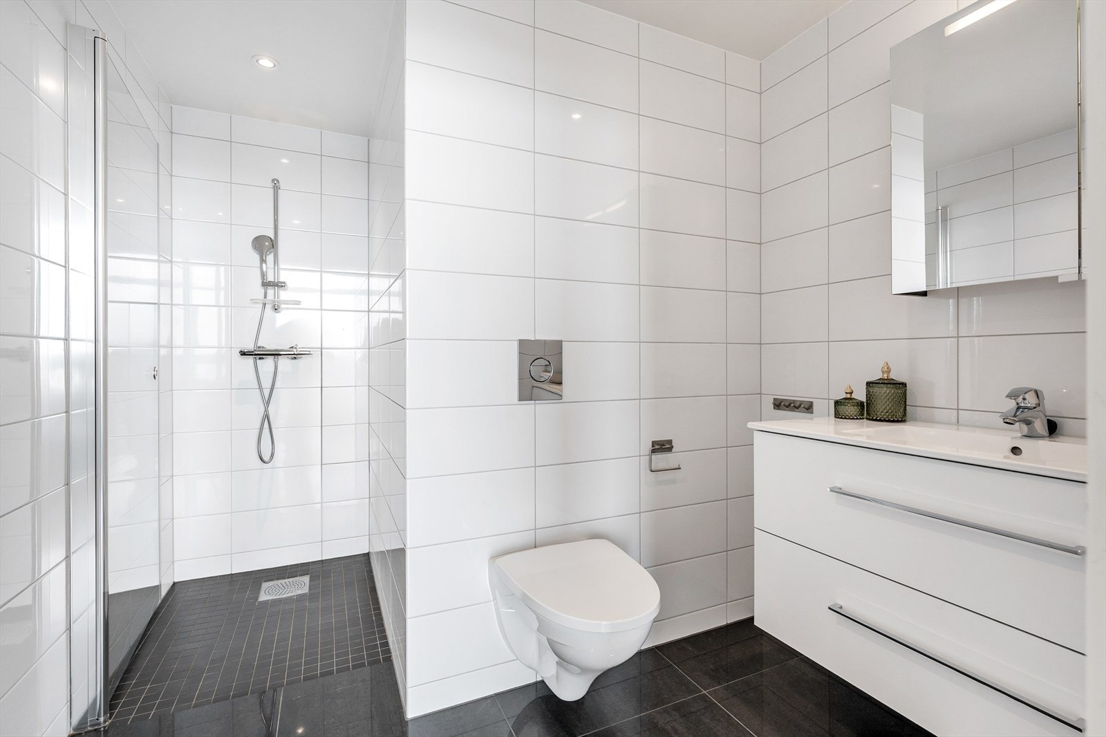 Flott baderom med fliser på gulv og vegger. Rommet er innredet med benkeskap med servant, blandebatteri som ble oppgradert i 2023, dusjnisje med glassdør og vegghengt toalett. Galleribilde