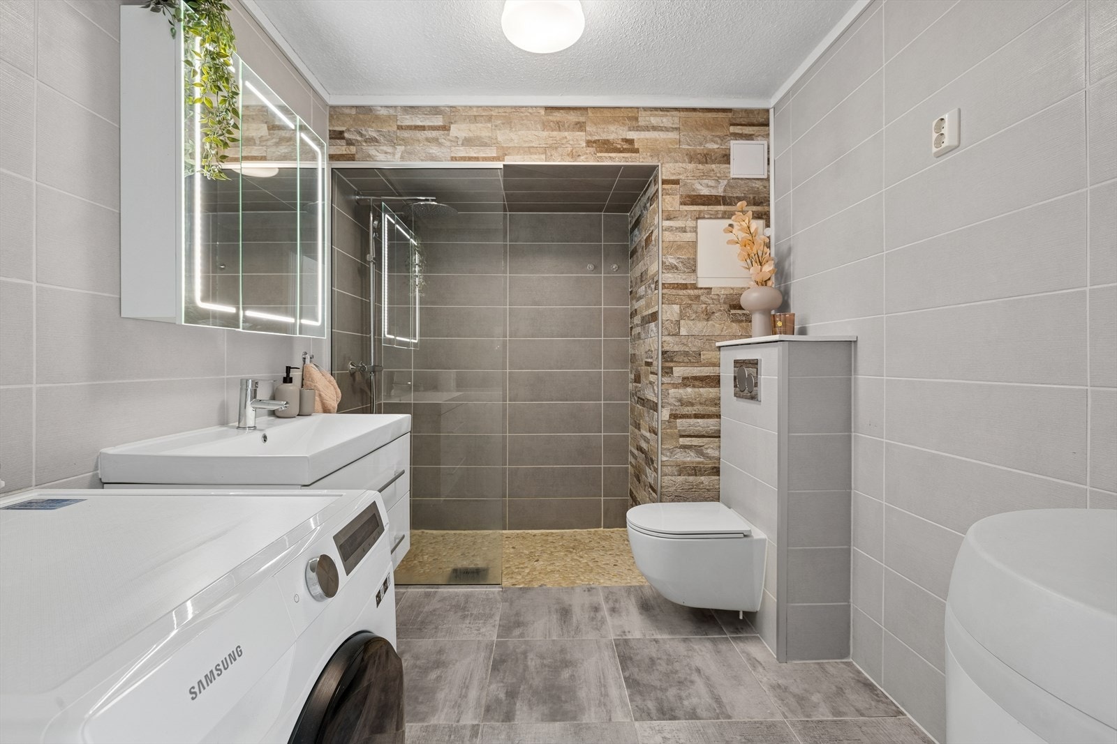 Badet fungerer som kombinert bad og vaskerom med fliser på gulv og vegger. Det er behagelige varmekabler for økt komfort. Galleribilde