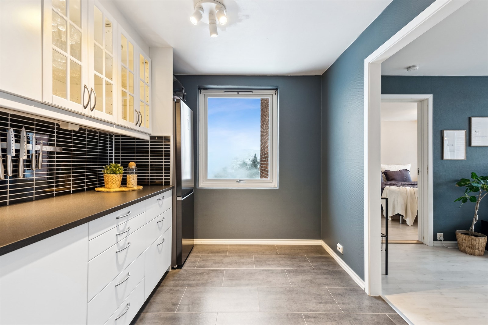 Delvis åpen løsning mellom stue og kjøkken. Det er lys i og under overskap, samt naturlig lys fra vindu. Galleribilde