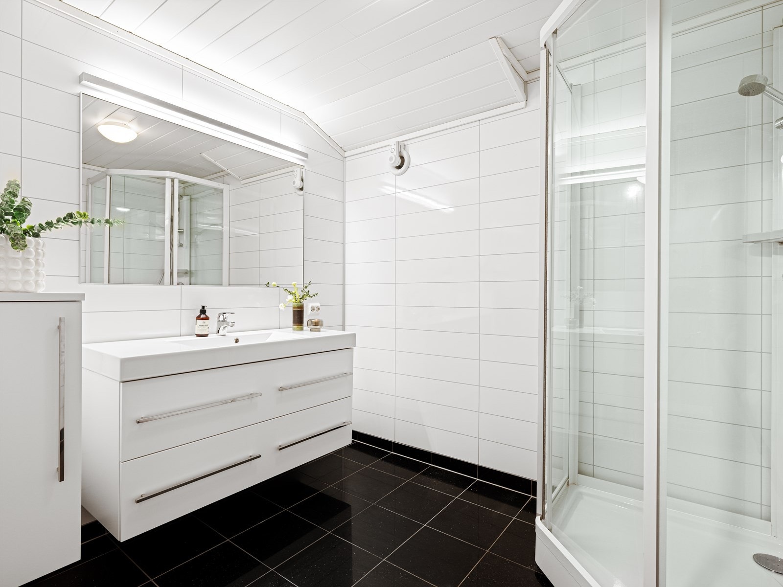 Stort bad med fliser på gulv og vegg. Dusjkabinett, vask i seksjon og speil med belysning. Vegghengt skap til lagring. Galleribilde