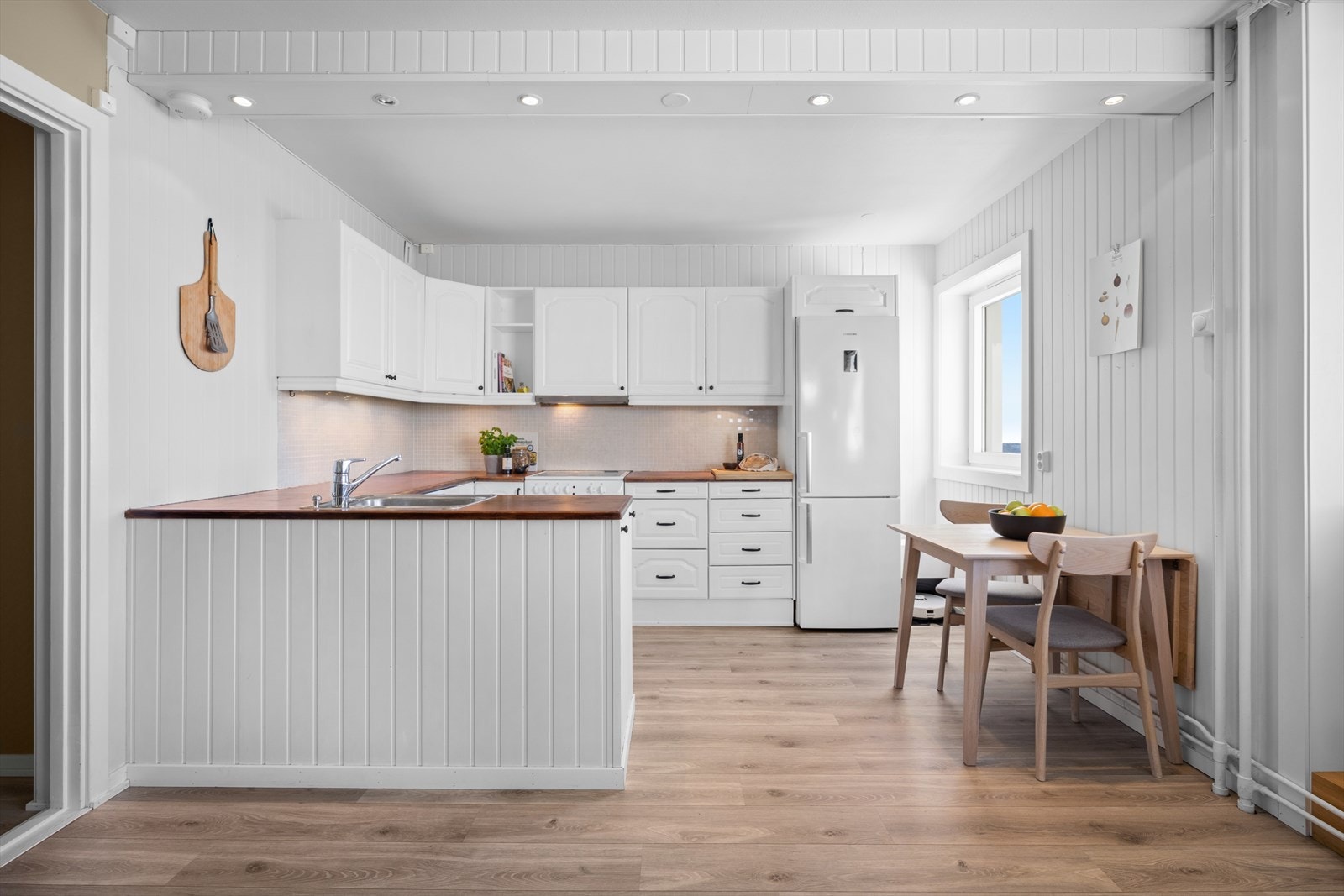 Åpen kjøkkenløsning med en tidløs innredning, profilerte fronter og heltre benkeplate med nedfelt kum i rustfritt stål. Galleribilde