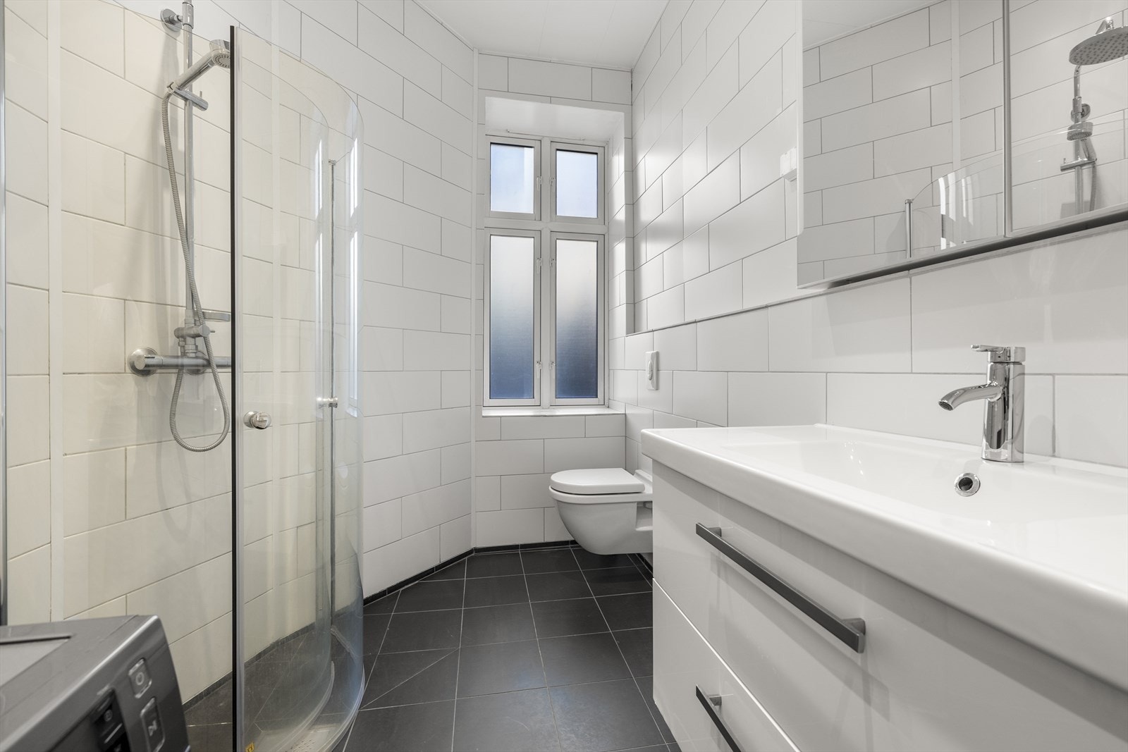 Her finner du en dusjløsning med innfellbare glassdører, stilren baderomsinnredning med nedfelt servant og speilskap, vegghengt toalett og opplegg for vaskemaskin. Galleribilde