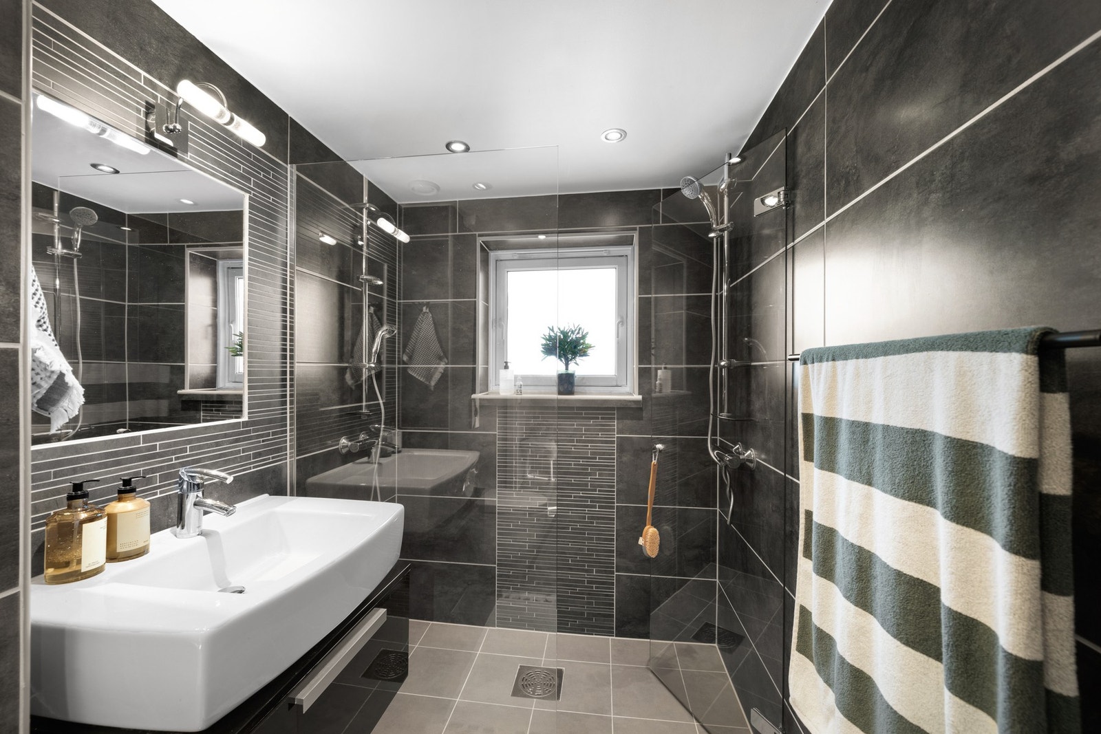 Dusjsonen har innfellbare glassdører og dobbel dusjløsning for en praktisk og luksuriøs opplevelse. Videre finner du et vegghengt toalett, servantskap med oppbevaringsplass, speil med belysning og opplegg for vaskemaskin. Galleribilde