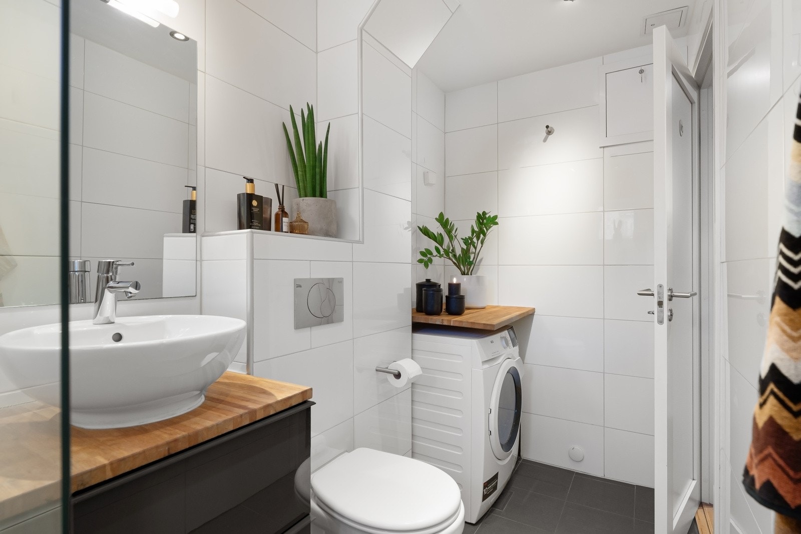 Stilren baderomsinnredning som harmonerer med gulvflisene, samt en elegant servant med tilhørende speil. De store hvite veggflisene kombinert med mørke gulvfliser gir badet et moderne og delikat uttrykk. Galleribilde