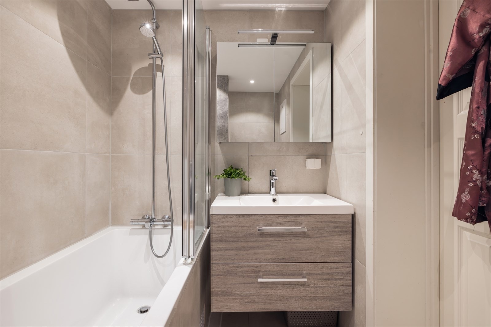 Innredningen består av servant i skuffeseksjon med 1-greps blandebatteri speilskap med lys, vegghengt wc og badekar med glassdører og regndusj Galleribilde