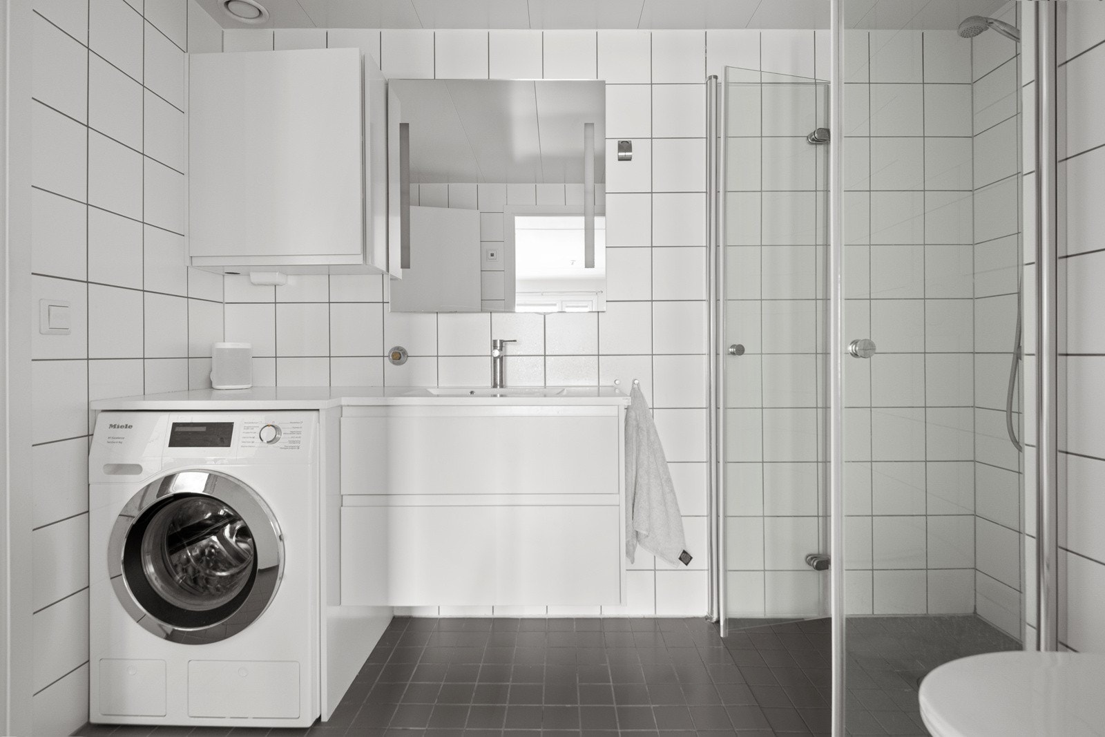 Moderne baderomsinnredning med skuffer og heldekkende servant med plass og opplegg for vaskemaskin under. Over er det montert vegghengt skap og stort speil med integrert belysning. Galleribilde