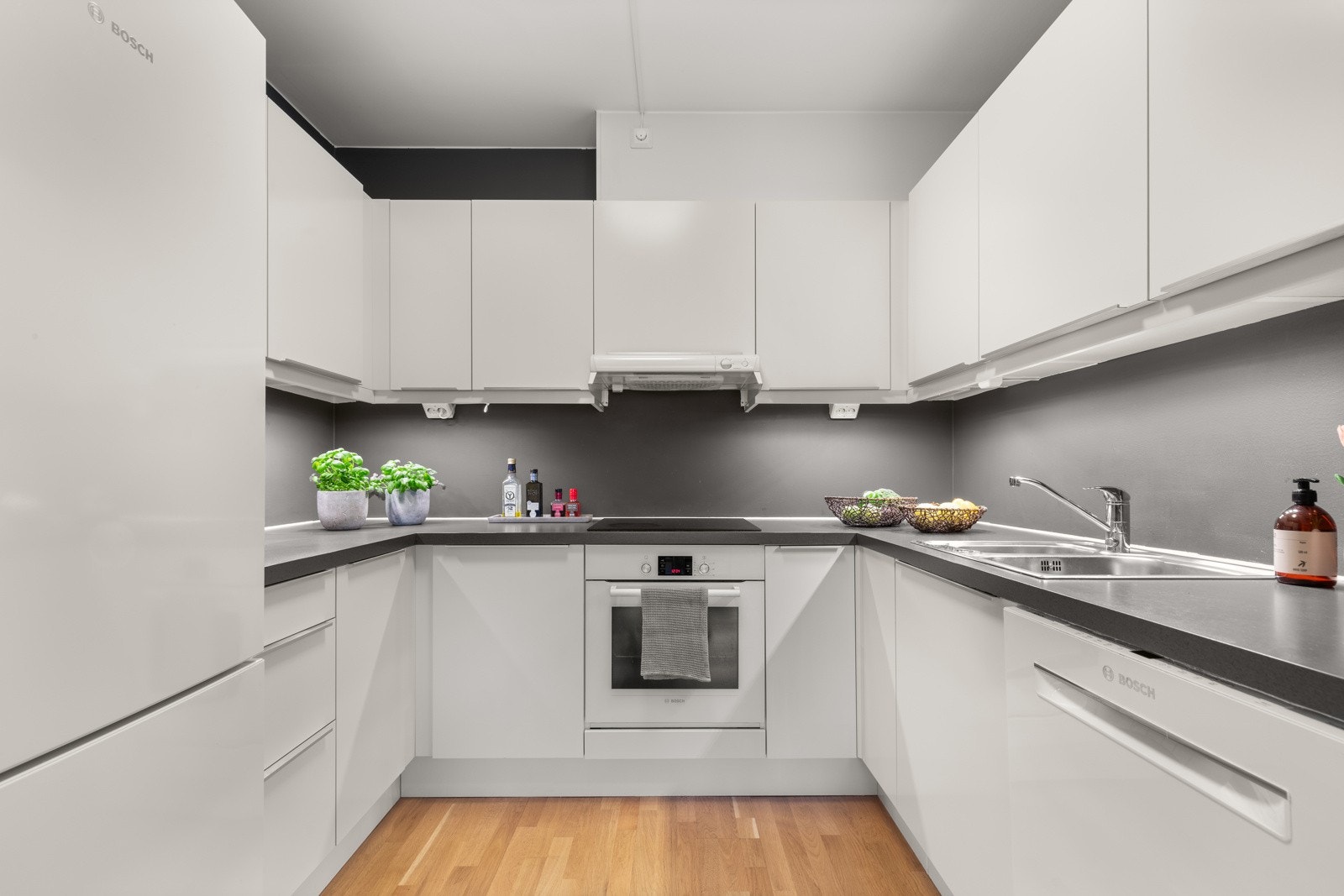 Kjøkken fra HTH med hvite, slette fronter, malt backsplash og benkeplate i laminat. Kjøkkenløsningen er plassert i en praktisk U-formet nisje, som gir gode arbeidsforhold for matlaging. Galleribilde