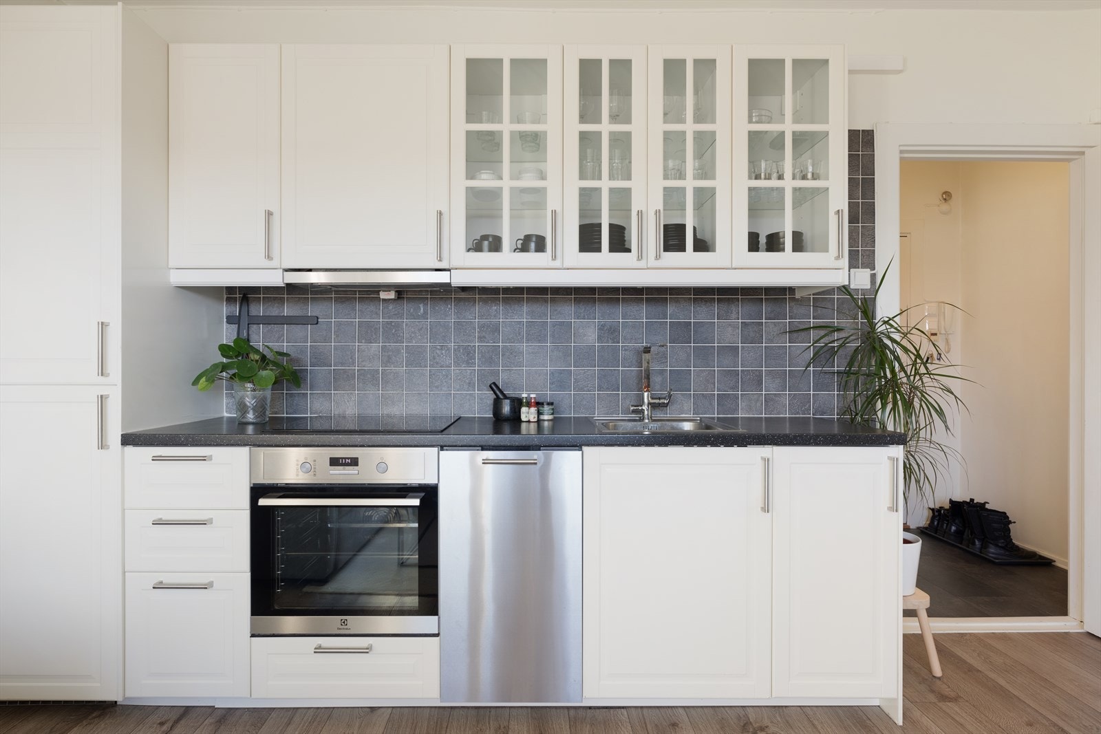 Langs den ene langveggen er den stilrene kjøkkeninnredningen plassert. Innredningen er fra 2016 med hvite fronter og laminat benkeplate med oppvaskkum. Det er godt med oppbevaringsplass i skuffer og skap som gjør det enkelt å holde orden. Galleribilde