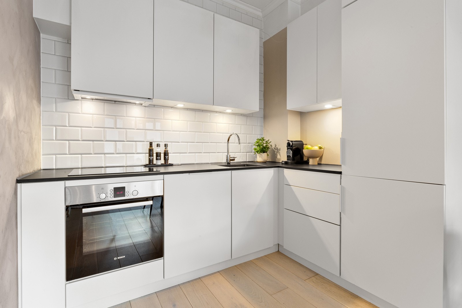 Det har slette skapfronter, laminerte benkeplater og flislagt backsplash. Hvite fliser i bakkant av benkeplaten og godt med belysning under overskapene Galleribilde