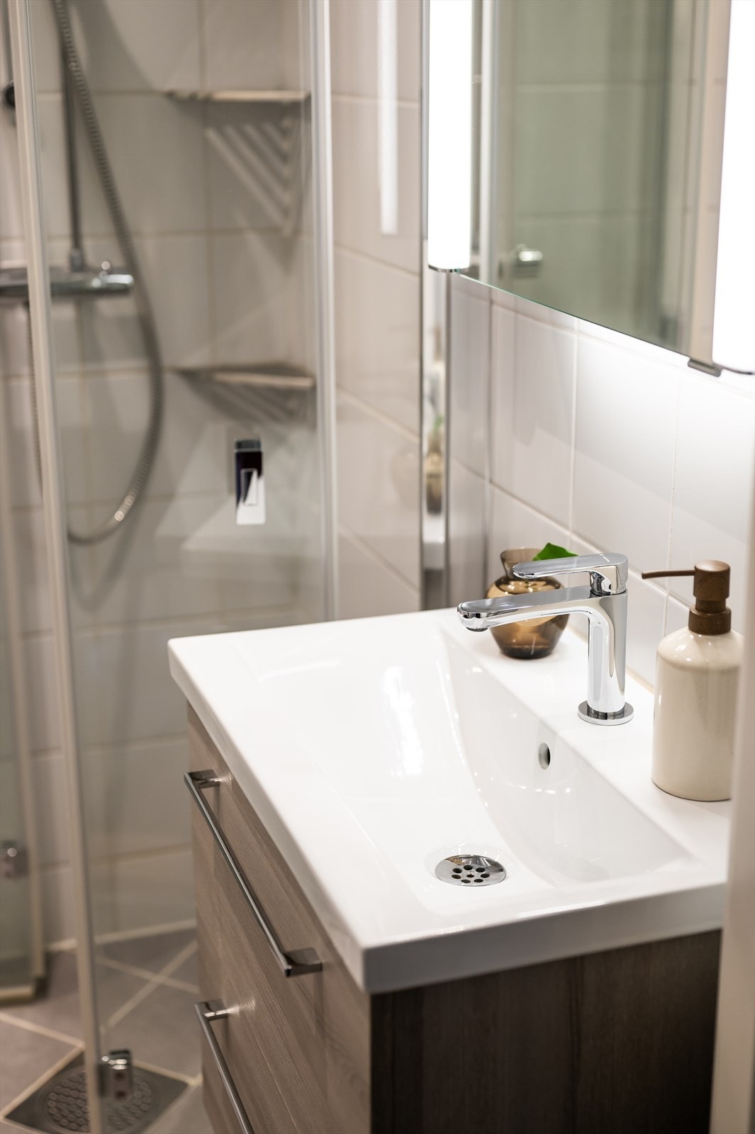 Videre har badet dusjhjørne med leddet glassdører og dusjarmatur tilkoblet veggmontert hånddusj, vegghengt toalett med innebygget sisterne og opplegg for vaskemaskin. Avtrekksventil plassert i himling. Galleribilde