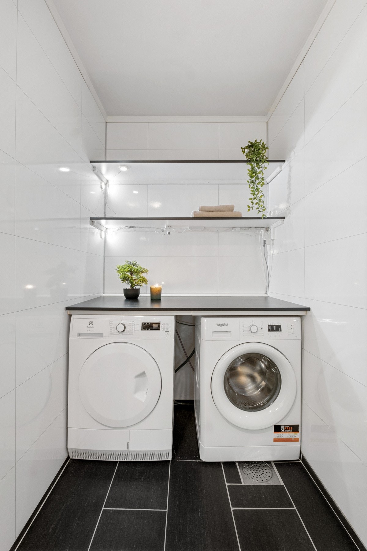 Vaskeromsdel med nisje for vaskemaskin og tørketrommel under en benkeplate. Over benken er det hyller på veggen. Galleribilde