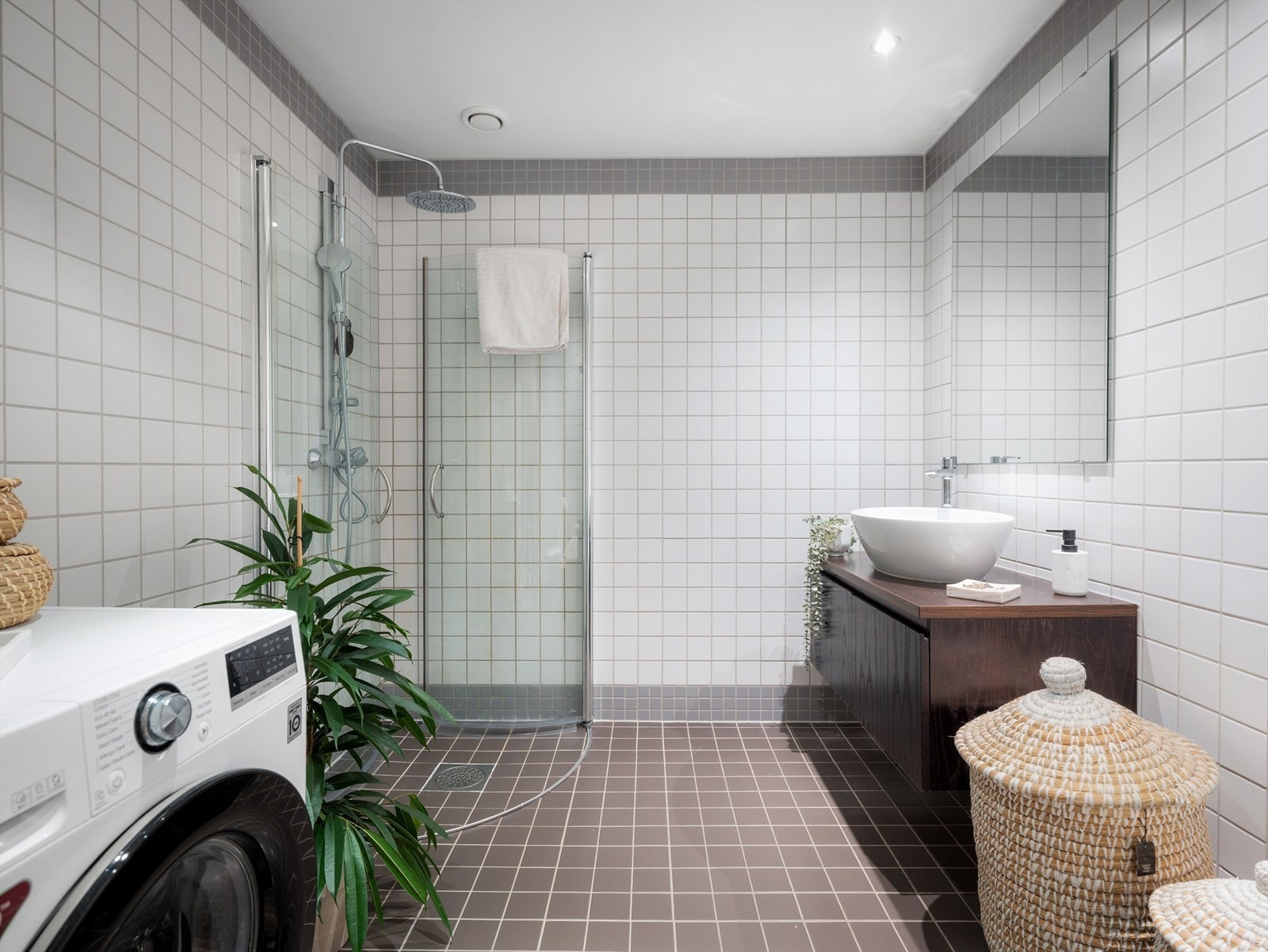 Flislagt bad som inneholder servant med underskap, dusjshjørne med innfeltbare glassdører, toalett og opplegg til vaskemaskin Galleribilde