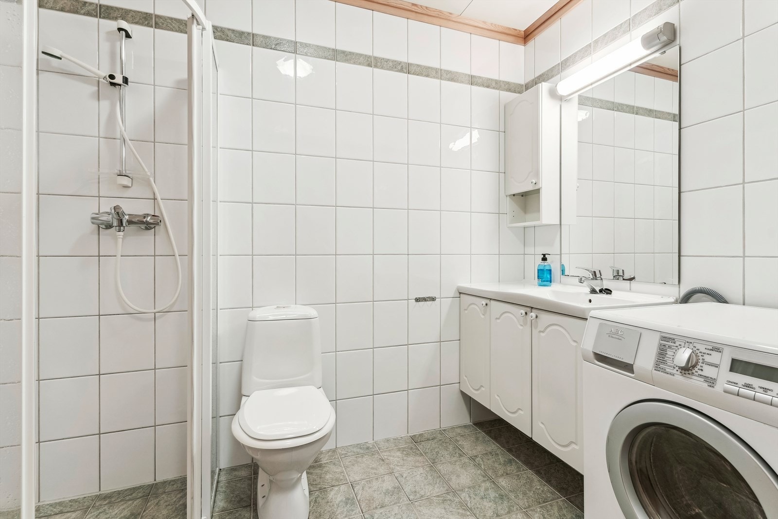 Baderom fra byggeår med dusj, toalett, servant med innredning og opplegg til vaskemaskin. Galleribilde