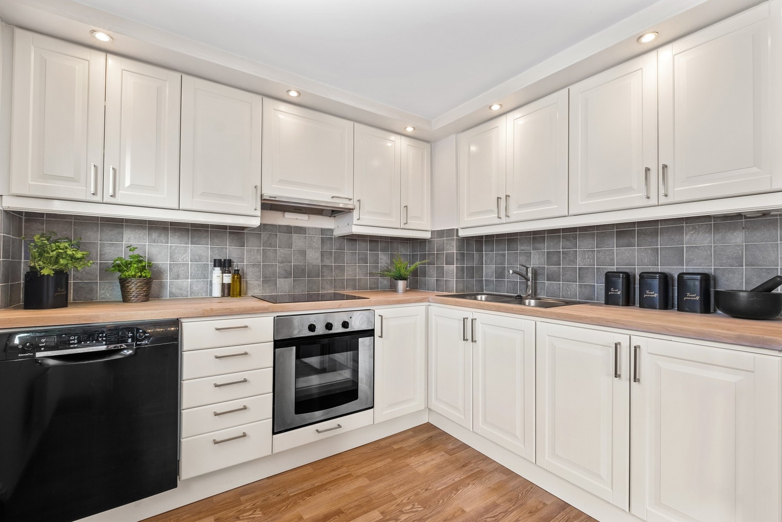 Glatte og malte fronter samt kjøkkenbenk av eik. Det er montert fliser og spotlys mellom benk og overskap, samt keramisk topp og ventilator over kokesonen. Komfyrvakt er montert. Galleribilde