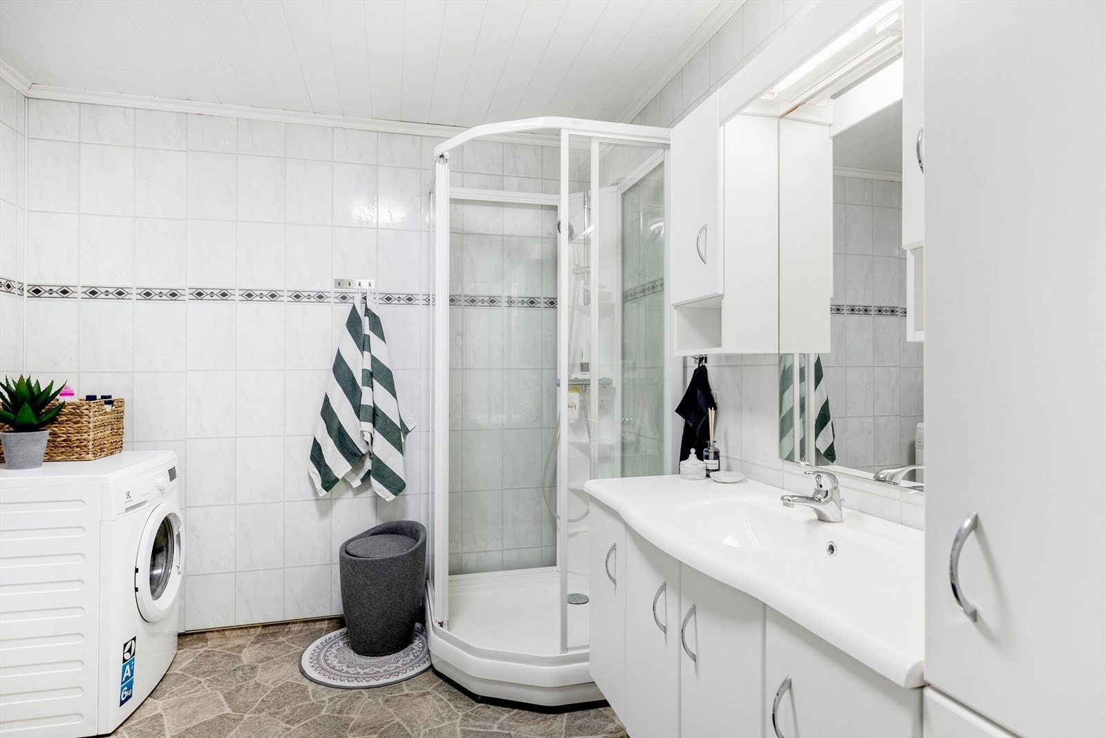 Baderommet er romslig og har belegg med varme i gulv, baderomsplater på vegger og MDF-panel i himling. Badet inneholder baderomsinnredning, dusjkabinett, vegghengt toalett og opplegg for vaskemaskin Galleribilde