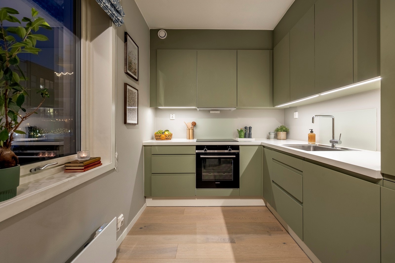 Kjøkkeninnredningen er designet i en stilren L-form langs to vegger, med malte, slette fronter på skap og skuffer som har praktisk gripekant. Galleribilde